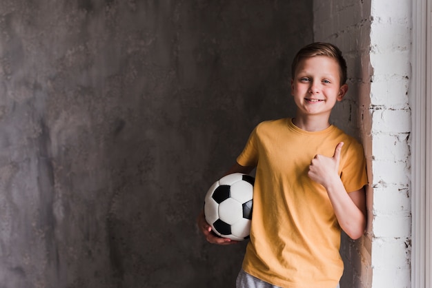 Foto retrato, de, um, menino sorridente, frente, parede concreta, segurando, bola futebol, mostrando, polegares cima