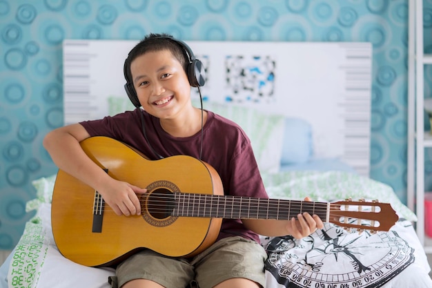 Retrato de um menino sorridente ouvindo música e tocando guitarra enquanto está sentado na cama em casa