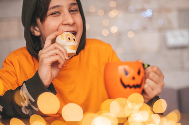 Retrato de um menino zoomer autêntico e sincero posando com uma xícara de chá e doces em casa, festa de Halloween