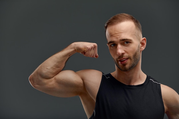 Retrato de um modelo de cara jovem bonito com penteado em um top preto na moda simulado com um corpo musculoso saudável mostrando seus músculos bíceps em um fundo cinza