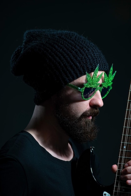 Retrato de um músico barbudo usando óculos escuros de maconha tocando emocionalmente a guitarra elétrica em um fundo escuro