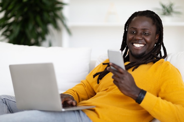 Retrato de um negro sorridente usando laptop e lendo mensagem no smartphone