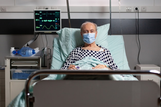Retrato de um paciente idoso doente com máscara cirúrgica, descansando em uma cama de hospital