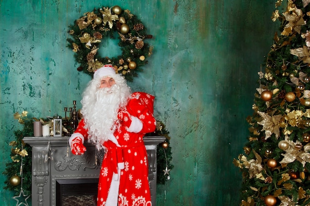 Retrato de um Papai Noel emocional com saco de presentes perto da lareira de Natal na sala decorada com árvore de Natal. Papai Noel vem e traz presentes à noite. Copie o espaço para o site