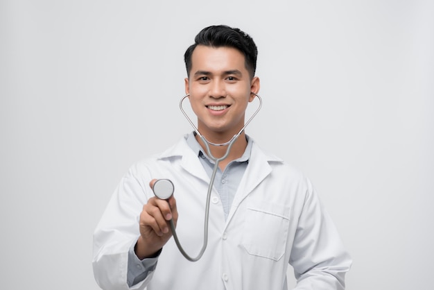 Retrato de um simpático médico bonito vestido de uniforme segurando um estetoscópio e olhando para a câmera isolada sobre fundo branco