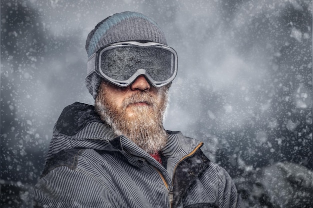 Retrato de um snowboarder ruiva com uma barba cheia em um chapéu de inverno e óculos de proteção vestido com um casaco de snowboard posando no contexto das montanhas.