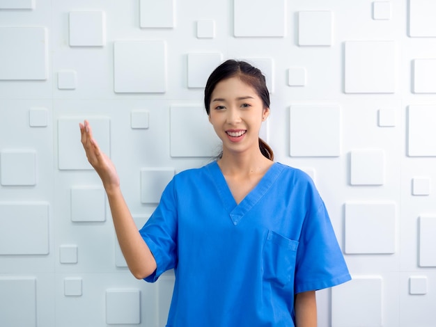 Retrato de um sorriso amigável e alegre, uma mulher asiática confiante, médica ou enfermeira, fazendo um gesto explicativo com a mão no fundo branco, olhando para a câmera, profissional de saúde em uniforme azul