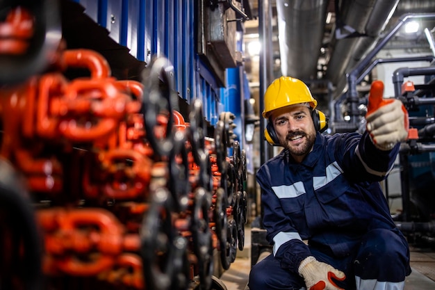 Retrato de um trabalhador alegre da usina de aquecimento, parado ao lado de válvulas de tubos e segurando os polegares para cima