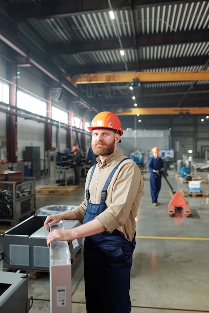 Retrato de um trabalhador manual confiante de macacão e capacete segurando um equipamento de metal em uma loja de fábrica contemporânea