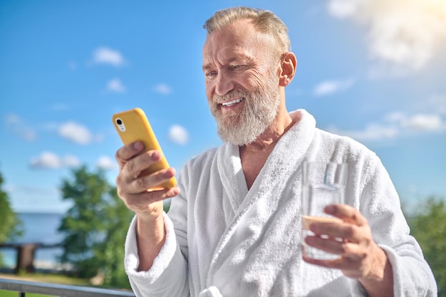 Retrato de um turista satisfeito com um copo de água e o celular em pé na varanda