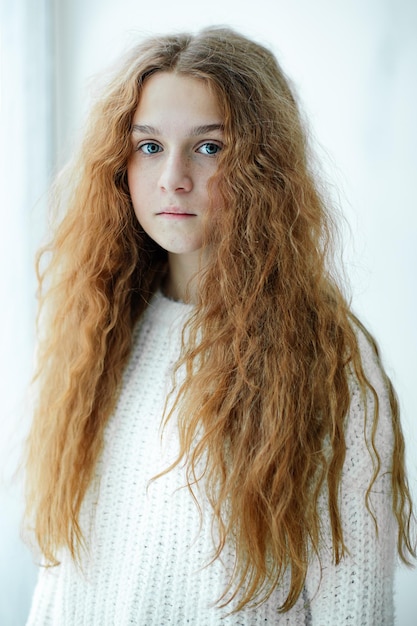 Retrato de uma adolescente de olhos azuis com luxuosos cabelos ruivos longos e ondulados em pé perto da janela à luz do dia de inverno. Lindas sardas. Mundo interior. Cabelo desgrenhado fofo. Beleza natural. Luz natural.