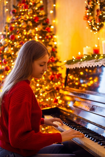 Retrato de uma adolescente de suéter vermelho tocando piano na sala de estar de natal decorada atmosfera acolhedora e aconchegante de celebrar o natal em casa belas decorações ao redor