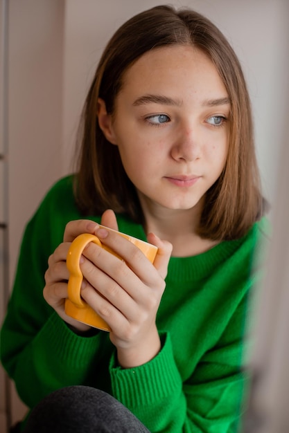 Retrato de uma adolescente em uma camisola verde com uma caneca laranja nas mãos dela. menina bebe chá ou café sentado no parapeito da janela e olhando pela janela. sonhos, romance. Foto de alta qualidade