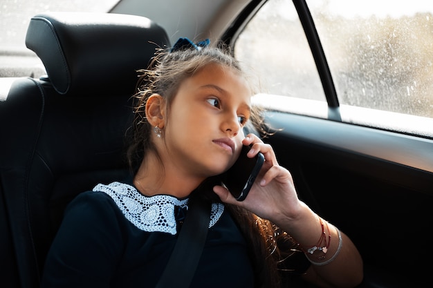 Retrato de uma adolescente falando em smartphone no carro, olhando pela janela. Vestindo forma escolar.