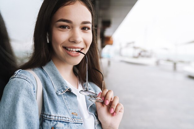 Retrato de uma adolescente morena atraente usando jaqueta jeans, segurando óculos e sorrindo para a câmera enquanto caminha ao ar livre