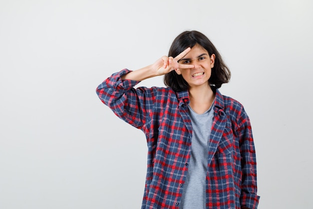 Retrato de uma adolescente mostrando o V-sign no olho com roupas casuais e uma alegre vista frontal