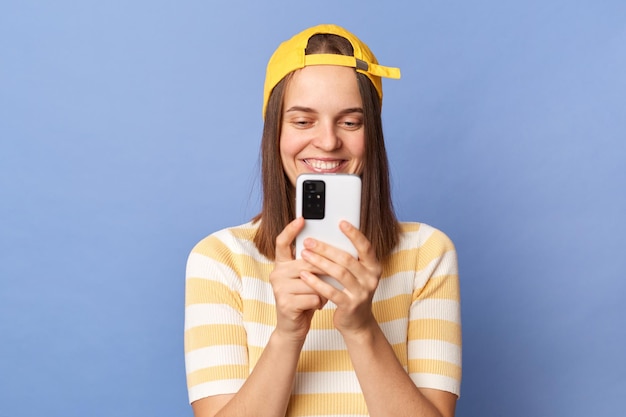 Retrato de uma adolescente sorridente vestindo camiseta listrada e boné de beisebol em pé isolado sobre fundo azul segurando smartphone conversando com amigos fazendo foto