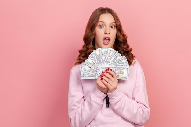 Retrato de uma adolescente sortuda com capuz surpreso ao segurar um monte de notas de dólar na mão, chocado com a grande vitória na loteria, economia financeira. tiro de estúdio interior isolado no fundo rosa