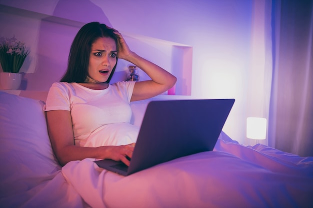 Retrato de uma adorável encantadora mulher nervosa e intrigada deitada na cama usando um laptop