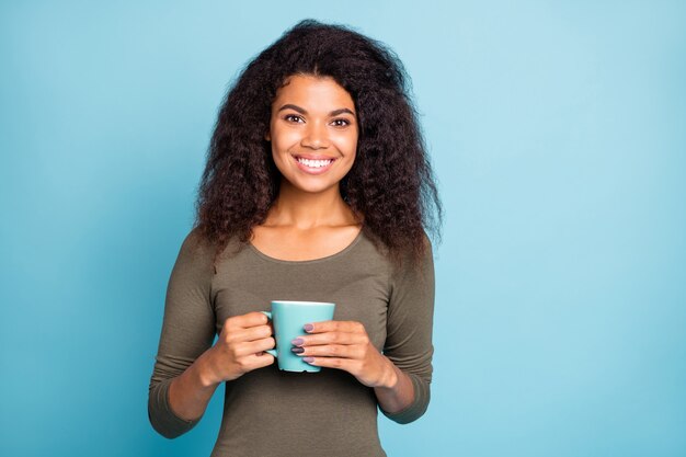 Retrato de uma adorável garota mulata positiva segurando uma xícara de café com cacau e sentindo-se contente nos finais de semana. Use roupa casual isolada sobre a parede azul