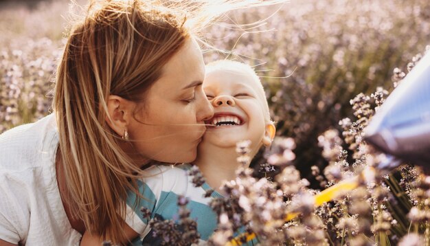 Retrato de uma adorável jovem mãe beijando seu filho enquanto ele está rindo com os olhos fechados, por do sol em um campo de flores.