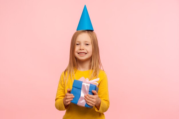 Retrato de uma adorável menina feliz com cone de festa engraçada na cabeça, segurando a caixa de presente e sorrindo alegremente para a câmera, animado com o presente de aniversário. foto de estúdio interno isolada em fundo rosa