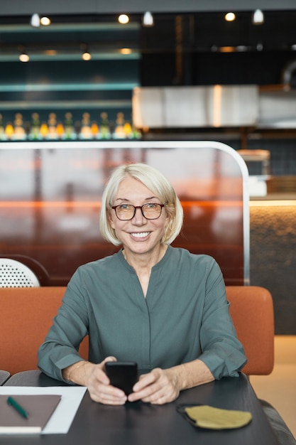 Retrato de uma alegre empresária caucasiana de cabelos loiros em óculos, segurando um smartphone e sorrindo para a câmera em um café moderno