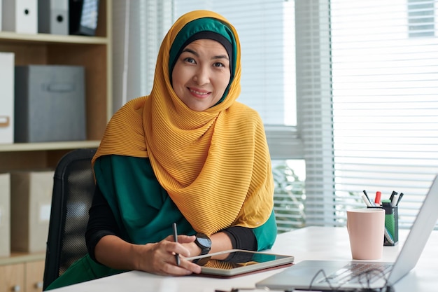 Retrato de uma alegre empresária muçulmana preenchendo um formulário em um computador tablet