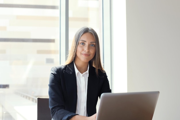 Retrato de uma alegre jovem empresária sentado à mesa no escritório e olhando para a câmera.