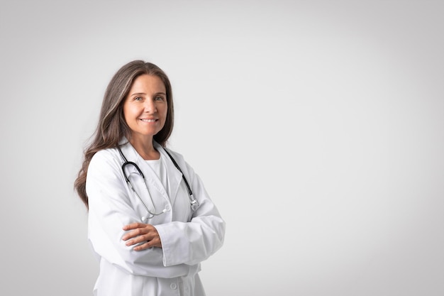 Retrato de uma alegre médica europeia sênior em jaleco branco com estetoscópio em pé com espaço de cópia de braços cruzados