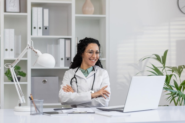 Retrato de uma alegre médica hispânica usando óculos sentada em seu escritório da clínica com um