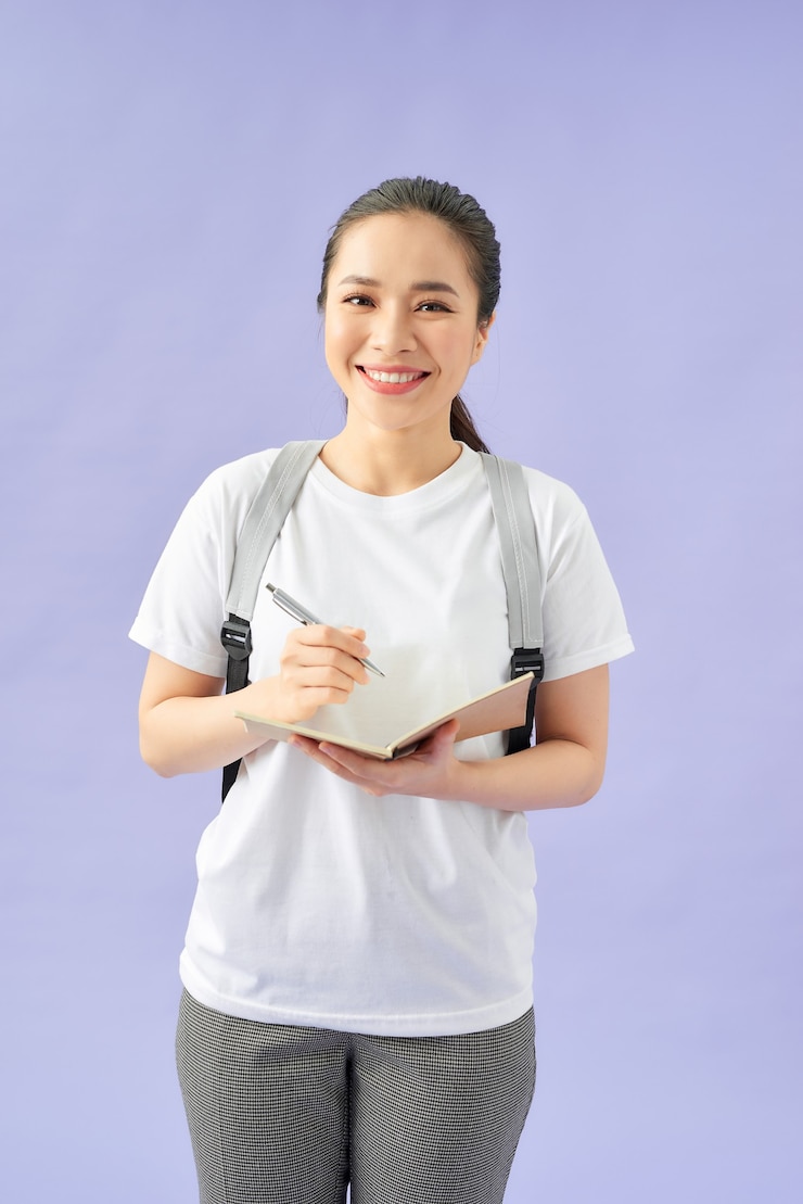 Foto retrato de uma aluna feliz e casual com uma mochila escrevendo em um bloco de notas em pé com livros isolados sobre fundo roxo