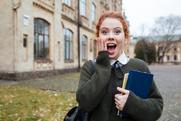 Retrato de uma aluna ruiva atônita segurando livros fora do campus da universidade