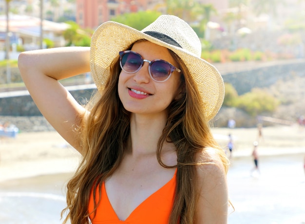 Retrato de uma atraente garota sorridente com cabelo comprido, posando na praia. Ela usa biquíni laranja, chapéu de palha e óculos escuros.