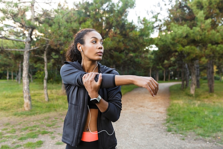 Foto retrato de uma atraente mulher afro-americana de 20 anos, vestindo um agasalho esportivo preto, fazendo exercícios e esticando o corpo no parque verde
