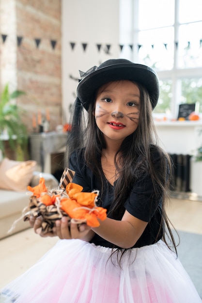 Retrato de uma bela garota asiática com maquiagem de gatinho e chapéu preto segurando uma pilha de doces de Halloween