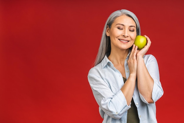 Retrato de uma bela idosa madura asiática idosa segurando uma maçã sorrindo isolada sobre fundo vermelho Uma maçã por dia mantém um médico afastado