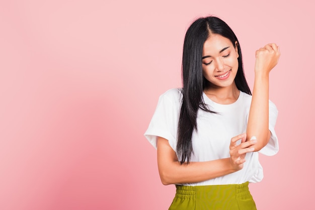 Retrato de uma bela jovem asiática aplicando loção creme hidratante cosmético na pele do cotovelo, foto de estúdio isolada em fundo rosa, conceito de cuidados com o corpo médico e higiênico