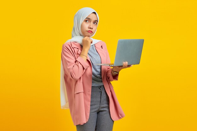 Retrato de uma bela jovem asiática segurando um laptop e, pensativa, segurando o queixo isolado sobre um fundo amarelo