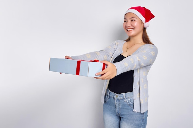 Retrato de uma bela jovem asiática sorridente vestindo um suéter azul com um chapéu de Papai Noel dando uma caixa de presente isolada no fundo branco do estúdio Feliz Natal Conceito