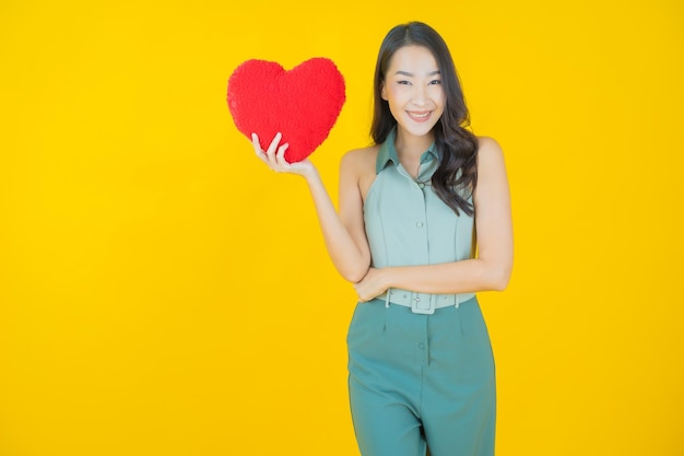 Retrato de uma bela jovem asiática sorrindo com formato de almofada em forma de coração na parede amarela