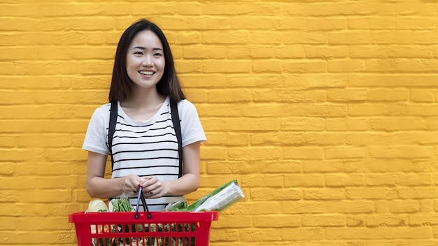 Foto retrato de uma bela jovem asiática sorrindo com uma cesta de compras do supermercado em uma cor amarela