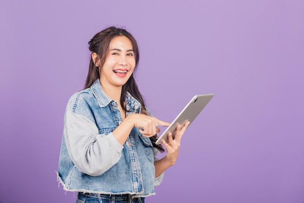 Retrato de uma bela jovem asiática sorrindo usando computador tablet, tela sensível ao toque de uma adolescente feminina de estilo de vida feliz no tablet pc digital, tiro de estúdio isolado em fundo roxo