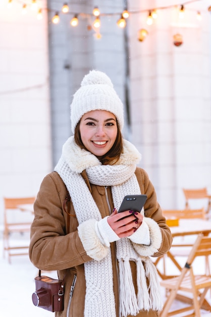 retrato de uma bela jovem com chapéu e lenço, caminhando ao ar livre na neve do inverno, bebendo café usando telefone celular.