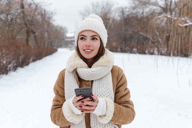 retrato de uma bela jovem com chapéu e lenço, caminhando ao ar livre na neve do inverno, usando telefone celular.