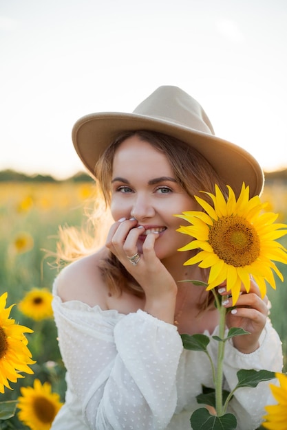 Retrato de uma bela jovem de 33 anos com chapéu no campo de girassol ao pôr do sol Modelo feliz em vestido branco na noite de verão na natureza Quente