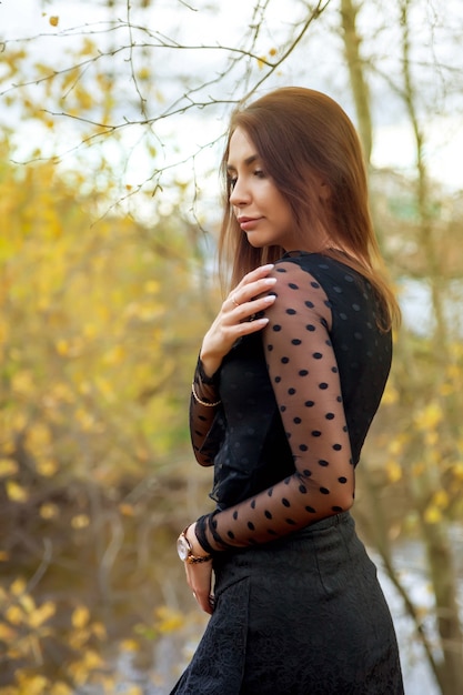 Retrato de uma bela jovem de aparência eslava em um vestido escuro no outono, em pé contra o fundo de um parque de outono e um lago com água