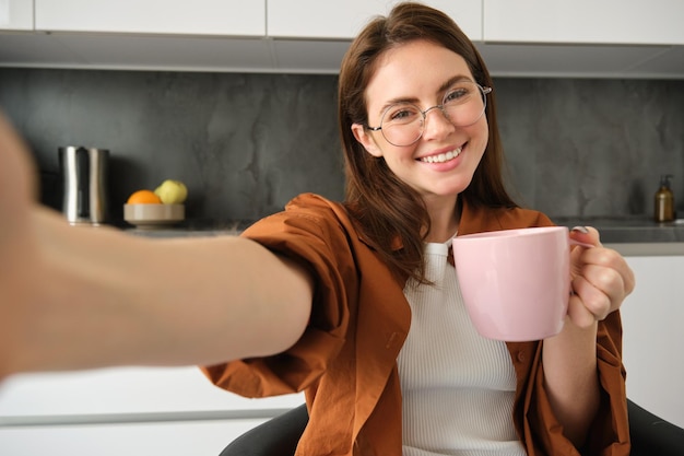 Retrato de uma bela jovem de óculos tirando uma selfie na cozinha com uma xícara de café da manhã