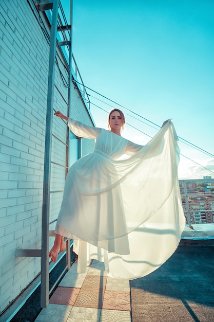 Retrato de uma bela jovem de vestido branco longo em pé no telhado do prédio com paisagem urbana