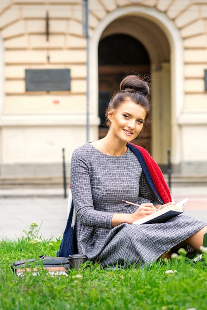 Retrato de uma bela jovem e sorridente estudante estudando com um livro na grama perto do prédio da universidade